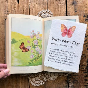 May include: A watercolor painting of a butterfly with pink and orange wings on a white background. The word "butterfly" is written below the image with a definition: "small creature with bright and colorful wings; symbol of hope and renewal; proof that change can be beautiful; spiritual messenger."