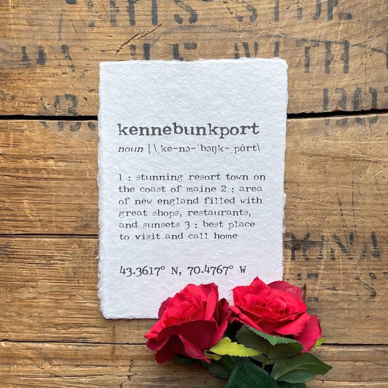 May include: A white paper print with the word "kennebunkport" defined as a stunning resort town on the coast of Maine. The print also includes the coordinates 43.3617&deg; N, 70.4767&deg; W.