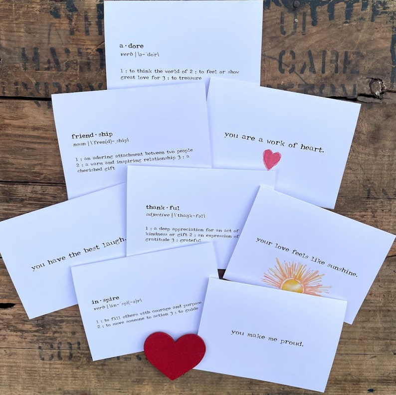 May include: A set of eight white cards with black text and red hearts. Each card features a different word and its definition. The words are "adore", "friendship", "thankful", "inspire", "you have the best laugh", "you are a work of heart", "your love feels like sunshine", and "you make me proud".