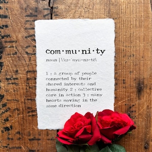 May include: A white paper with the word "community" printed in black, along with its pronunciation and definition. Two red roses rest at the bottom of the paper. The background is a rustic wooden surface.