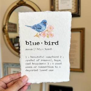 May include: A watercolor painting of a bluebird perched on a branch with pink blossoms. The image includes the text "blue bird" and a definition: "noun 'bli-, bərd 1: beautiful songbird 2: symbol of renewal, hope, and happiness 3: a good omen or connection to a departed loved one"