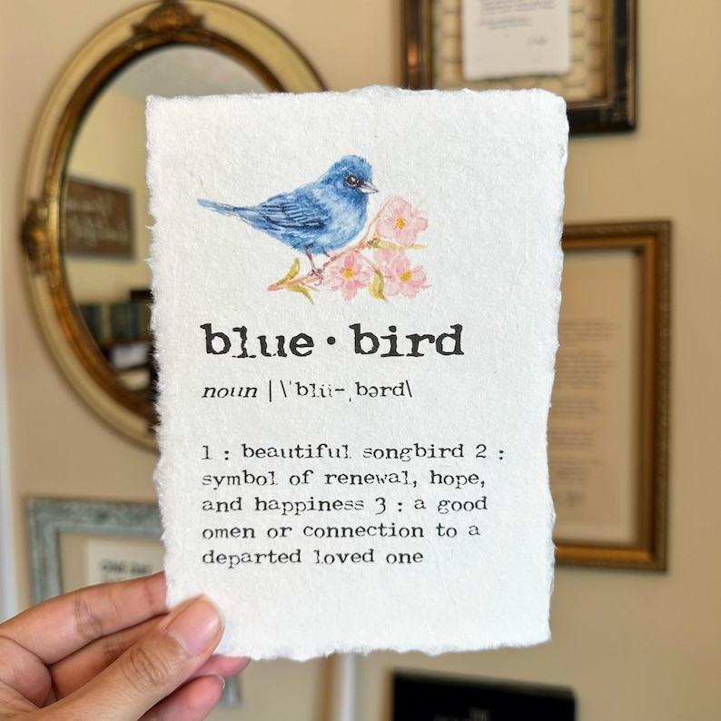 May include: A watercolor painting of a bluebird perched on a branch with pink blossoms. The image includes the text "blue bird" and a definition: "noun 'bli-, bərd 1: beautiful songbird 2: symbol of renewal, hope, and happiness 3: a good omen or connection to a departed loved one"