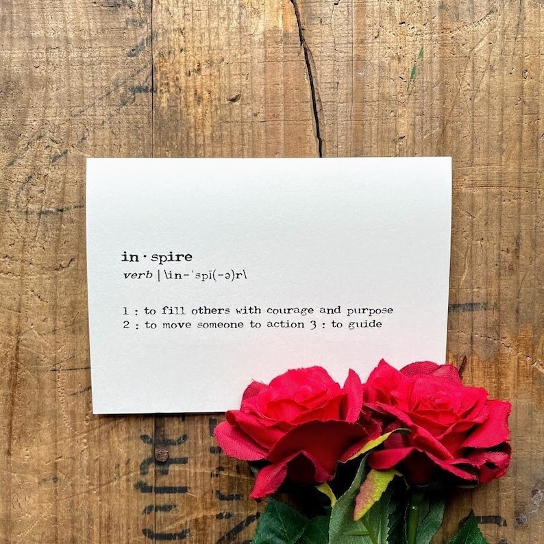 May include: A white card with the definition of the word "inspire" printed in black text. The card is sitting on a wooden surface with two red roses in the foreground.