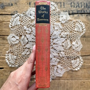 May include: A red hardcover book with gold lettering on the spine. The title is "The Works of Emerson".