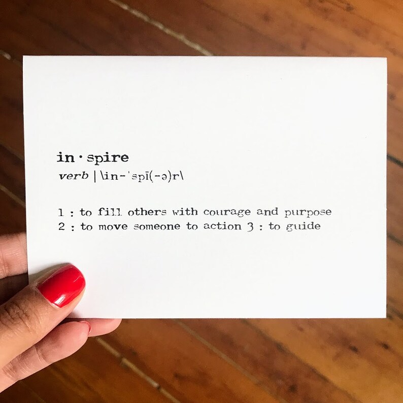May include: A white card with black text defining the word "inspire" as a verb. The definition includes three meanings: to fill others with courage and purpose, to move someone to action, and to guide.