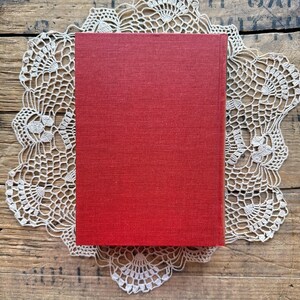 May include: A red hardcover book with a linen texture. The book is sitting on a white crocheted doily.