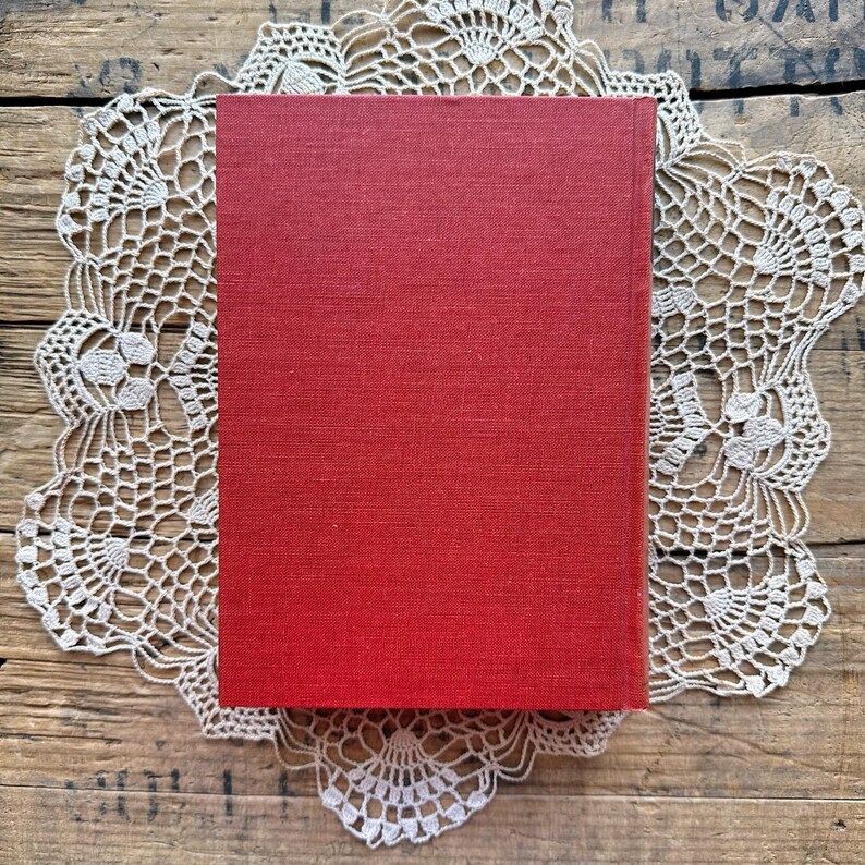 May include: A red hardcover book with a linen texture. The book is sitting on a white crocheted doily.