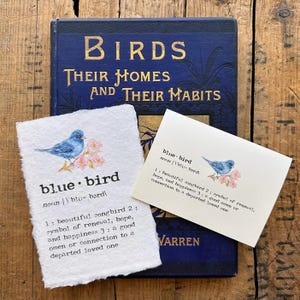 May include: Two cards with a bluebird illustration and the definition of the word "bluebird". The definition includes the meaning of the word as a symbol of renewal, hope, and happiness. The cards are on a wooden surface with a vintage book in the background.
