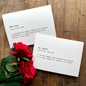 May include: Two white cards with black text defining the words "mentor" and "inspire". The cards are on a wooden surface with red roses in the foreground.