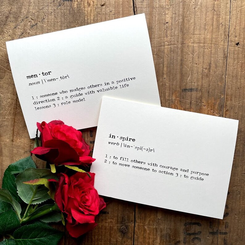 May include: Two white cards with black text defining the words "mentor" and "inspire". The cards are on a wooden surface with red roses in the foreground.