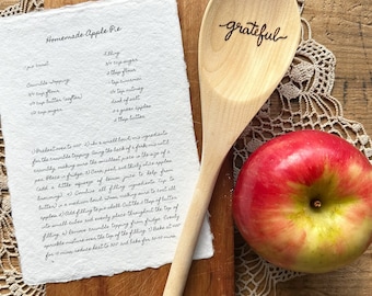 May include: A handwritten recipe for homemade apple pie on a piece of parchment paper with a wooden spoon that says "grateful" on it and a red apple next to it.