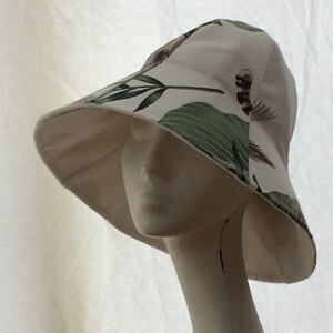 Puede incluir: Un sombrero de sol blanco con estampado de hojas tropicales verdes y marrones. El sombrero tiene un ala ancha y una corona suave y flexible.