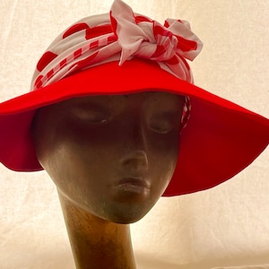 May include: A red sun hat with a wide brim. The hat is adorned with a white and red polka dot and striped fabric tied in a bow. The hat is displayed on a mannequin head.