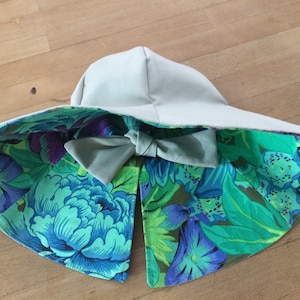 May include: A wide-brimmed sun hat with a floral print lining. The hat is made of a light beige fabric and has a large bow in the front. The floral print is a mix of green, blue, and purple.