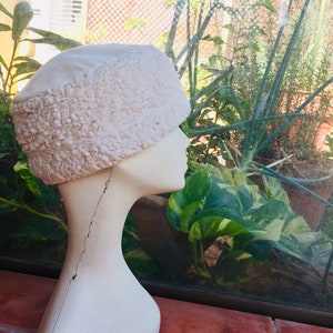 May include: A cream-colored hat with a textured, fuzzy brim and a smooth top, displayed on a mannequin head. The hat's design is reminiscent of a winter hat. The background features lush greenery and natural light.