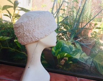 Puede incluir: Un sombrero color crema con un borde texturizado y difuso y una parte superior lisa, exhibido en una cabeza de maniquí. El diseño del sombrero recuerda a un sombrero de invierno. El fondo presenta una exuberante vegetación y luz natural.