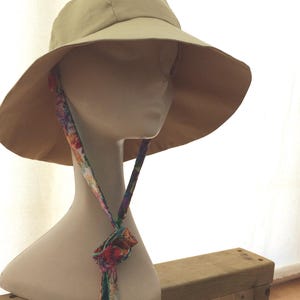 May include: A beige sun hat with a wide brim and a floral patterned chin strap. The hat is on a mannequin head.
