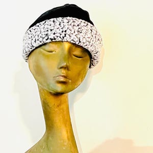 May include: A black beanie-style hat with a white and gray textured trim. The hat is displayed on a mannequin head with a bronze finish. The background is a plain, off-white color.