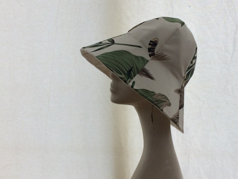 Puede incluir: Un sombrero de cubo blanco con un estampado de hojas tropicales verdes y marrones. El sombrero est&aacute; en una cabeza de maniqu&iacute; blanca.
