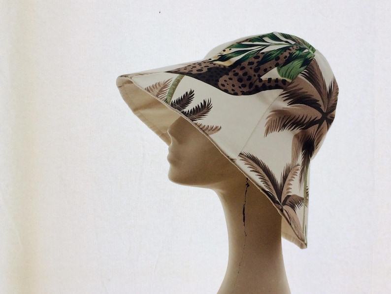 Puede incluir: Un sombrero de sol blanco con estampado tropical que presenta palmeras y un leopardo. El sombrero tiene un ala ancha y una corona redonda.