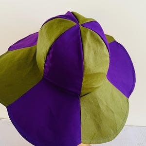 May include: A wide-brimmed sun hat with a unique design. The hat is made of two colours of fabric, a deep purple and a muted green. The hat has a rounded crown and a wide brim that is slightly curved.
