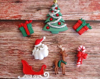 May include: A collection of Christmas-themed embellishments, including a green Christmas tree with red ornaments, a Santa Claus face, a reindeer with a green scarf, a red sleigh, two wrapped gifts, and a candy cane with a red bow. The items are arranged on a wooden surface.