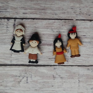 May include: Four small, decorative figurines representing historical figures. A woman in a black and white pilgrim outfit, a man in a black hat, a Native American woman, and a Native American man in a fringed outfit.