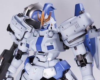Puede incluir: Un robot blanco y azul con una cresta dorada en la cabeza. El robot sostiene un arma negra en su mano derecha. El robot tiene el texto "TALLGEESE III" en su hombro izquierdo y "OZ-00MS" en su hombro derecho.