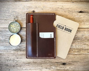 May include: A brown leather field book cover with a pen, card, and a "FIELD BOOK" card. A silver compass with a green face is to the left. The cover has white stitching.