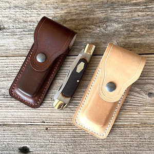 May include: A folding pocket knife with a dark brown handle and brass bolsters positioned between two leather sheaths. One sheath is dark brown with a metal snap closure, and the other is a light tan color with white stitching and a metal snap.