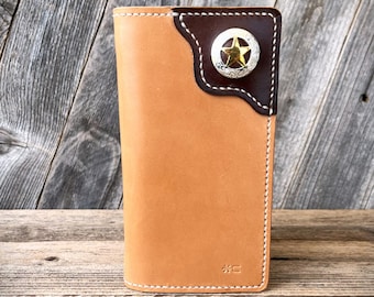 May include: A tan leather wallet with a dark brown accent on the top right corner. The accent features a silver-colored star-shaped emblem with a gold center. White stitching outlines the edges of the wallet.