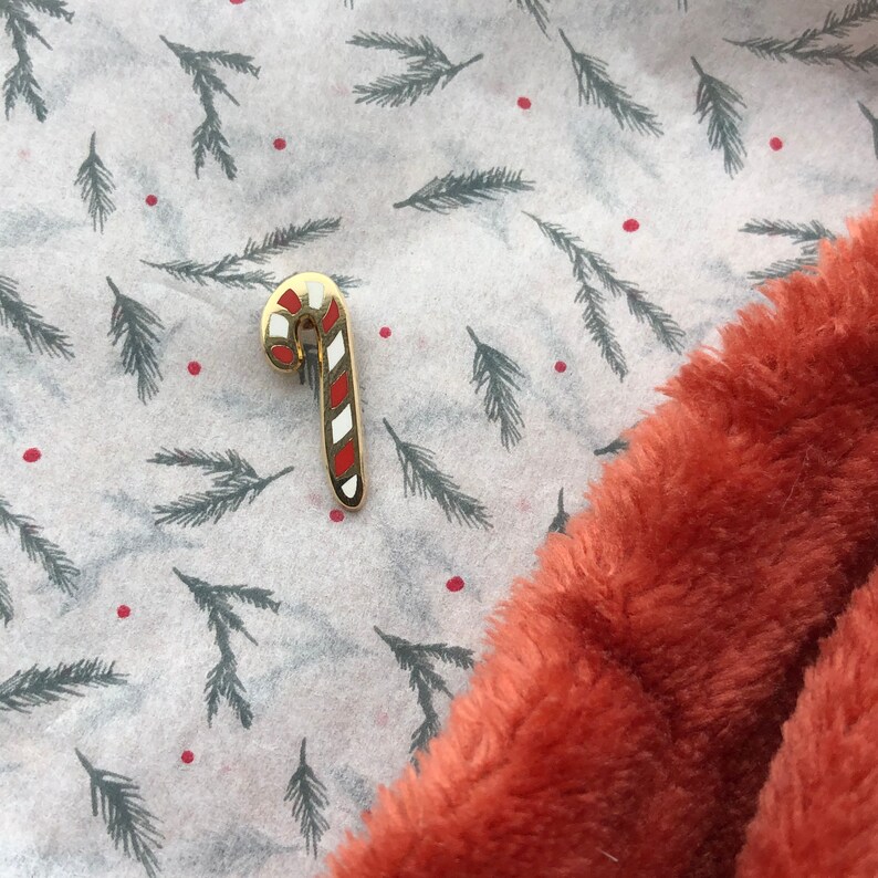 May include: A gold-colored candy cane pin with red and white stripes on a white background with a pattern of green pine branches and red dots.
