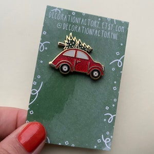 May include: A red car-shaped enamel pin with a Christmas tree on top. The pin has gold trim, black wheels, and white window details. The pin is on a green card with white decorative swirls and the text "DECORATIONFACTORY.ETSY.COM @DECORATIONFACTORYNL".