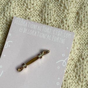 May include: A gold-colored pin shaped like a tool, possibly a spatula or similar, is displayed on a pink card. The pin has a light brown center and gold edges. The card has the text "DECORATIONFACTORY ETSY.COM @DECORATIONFACTORYNL".