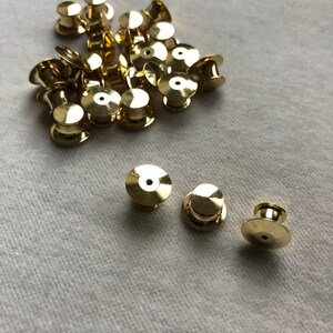 以下が含まれることがあります： ブローチ、ピン、その他のジュエリーに使用される、金色の金属製のピンバック（クラッチバックとも呼ばれます）。ピンバックは白い表面に散らばっています。