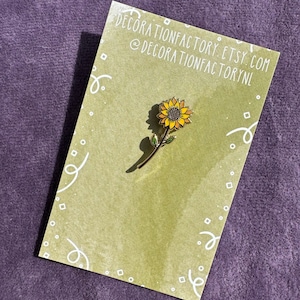 May include: A gold-colored enamel pin featuring a yellow sunflower with a green stem. The pin is on a green card with white polka dots and the text "decorationfactory.etsy.com @decorationfactorynl".