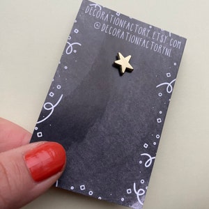May include: A gold star-shaped pin on a black card with white polka dots and a white squiggly line border. The card has the text "decorationfactory.etsy.com @decorationfactorynl" printed on it.
