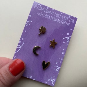 May include: Four gold-colored enamel pins on a purple background. The pins are shaped like a four-leaf clover, a star, a crescent moon, and a heart.