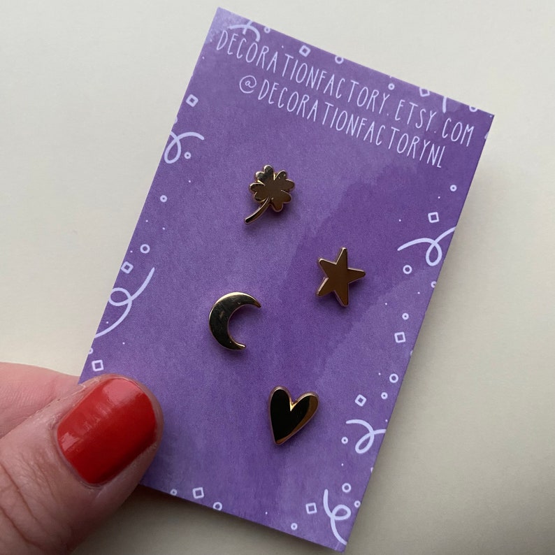 May include: Four gold-colored enamel pins on a purple background. The pins are shaped like a four-leaf clover, a star, a crescent moon, and a heart.