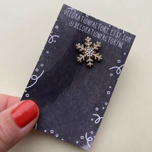 May include: A white snowflake enamel pin with a gold outline on a black background with white confetti. The pin is from Decorationfactory Etsy shop.