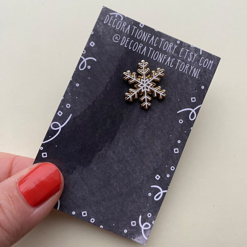 May include: A white snowflake enamel pin with a gold outline on a black background with white confetti. The pin is from Decorationfactory Etsy shop.