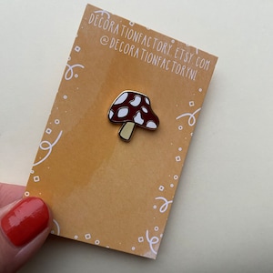 May include: A gold-coloured enamel pin featuring a red and white mushroom with white spots. The pin is on a brown card with the text "decorationfactory.etsy.com @decorationfactorynl".