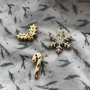 May include: Four gold-colored enamel pins with a Christmas theme. The pins feature a holly sprig, a snowflake, a candy cane, and a bell.
