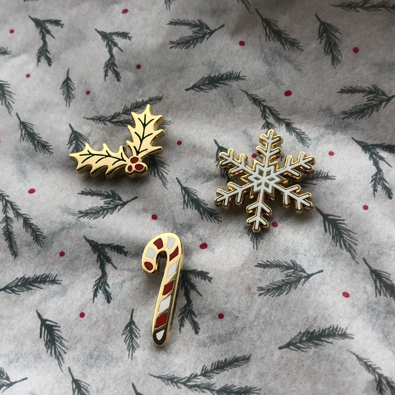 May include: Four gold-colored enamel pins with a Christmas theme. The pins feature a holly sprig, a snowflake, a candy cane, and a bell.