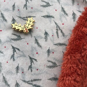 May include: A gold-colored holly leaf pin with red berries on a white background with a repeating pattern of dark green pine branches. The background is partially covered by a piece of fuzzy, rust-colored fabric.