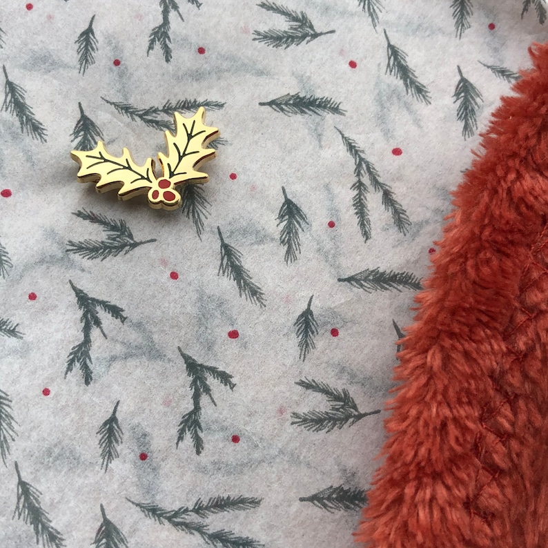 May include: A gold-colored holly leaf pin with red berries on a white background with a repeating pattern of dark green pine branches. The background is partially covered by a piece of fuzzy, rust-colored fabric.