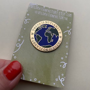 May include: A gold-coloured enamel pin with a blue and green world map design. The pin has the text "THERE IS NO PLANET B" in a circular pattern around the globe. The pin is on a green card with white text.