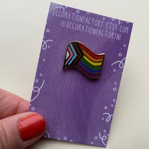Può includere: Uno spillo in smalto dorato con una bandiera arcobaleno con un motivo a strisce arcobaleno e un design a chevron con i colori della bandiera transgender. Lo spillo è su uno sfondo viola con pois bianchi e un bordo bianco.