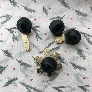 May include: Three gold-colored enamel pins with black metal backs. One pin is a snowflake design, one is a stick figure with a star on its head, and one is a stick figure with a star on its head and a star on its hand.