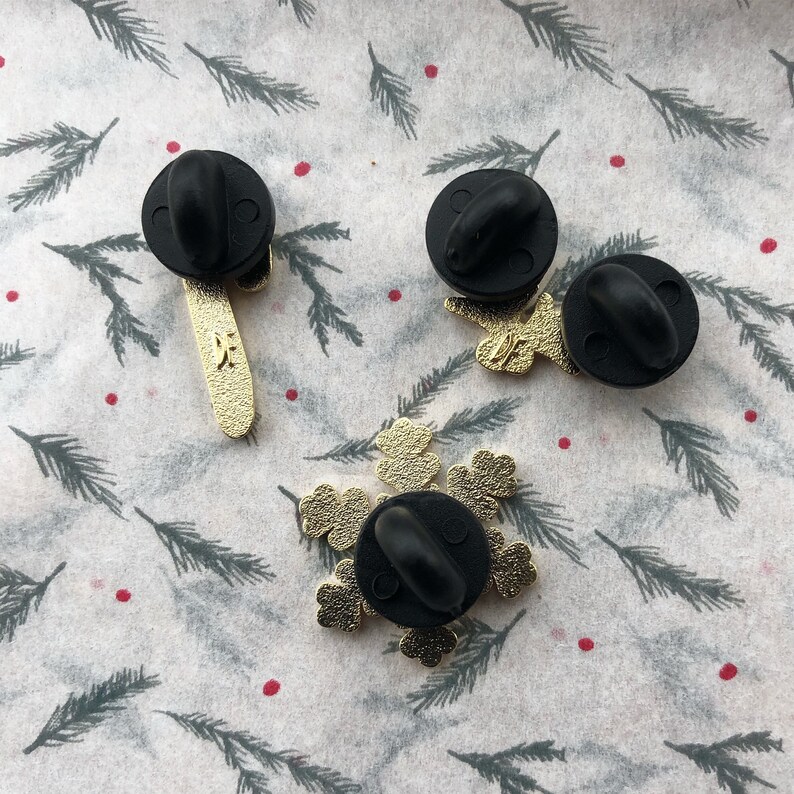 May include: Three gold-colored enamel pins with black metal backs. One pin is a snowflake design, one is a stick figure with a star on its head, and one is a stick figure with a star on its head and a star on its hand.